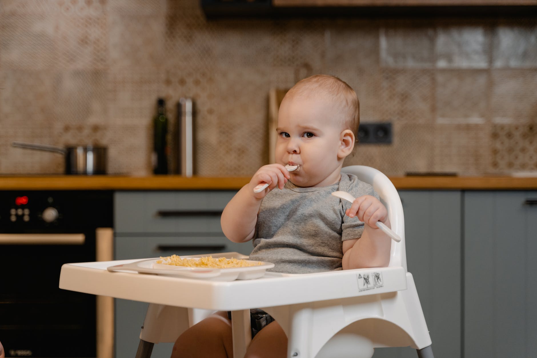a cut baby in gray shirt sitting on a high chair while eating food