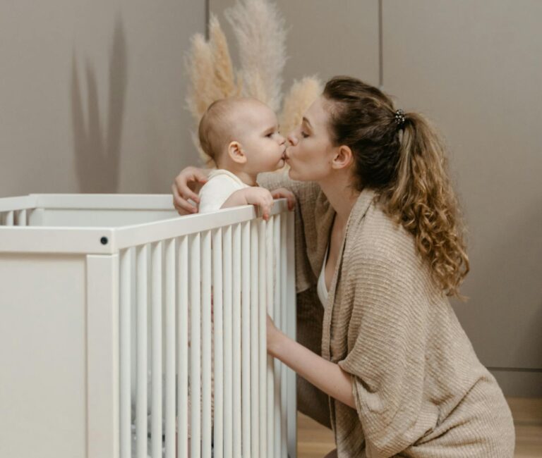 Room sharing and sleep training? Is it possible? A mother kisses her baby by the crib in a cozy nursery setting.